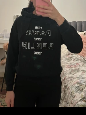 Svart hoodie med tryck Paris Berlin - Säljer en svart hoodie med vit text på bröstet där det står 'Paris 1989' och 'Berlin 1989'. Hoodien har huva och dragsko, samt ribbade muddar vid ärmar och nederkant. Perfekt för dig som gillar enkel streetstyle och vill ha något bekvämt och snyggt.