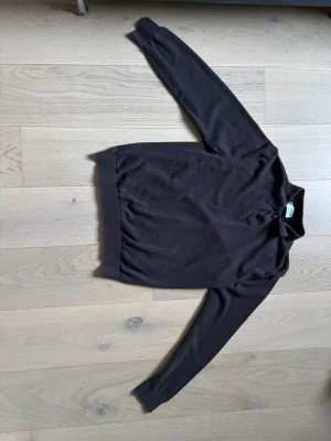 Mörkbrun pikétröja  - Klassisk mörkbrun pikétröja med lång ärm och ribbade muddar. Tröjan har krage och två knappar vid halsen för en stilren look. Perfekt för dig som gillar enkel och tidlös stil.