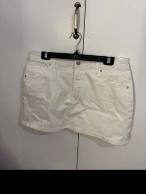 Vit kort jeanskjol med fickor - Snygg vit kort jeanskjol med klassisk femficksdesign och bälteshällor. Kjolen har knapp och dragkedja framtill samt två bakfickor. Perfekt till sommaren och enkel att matcha med olika toppar.