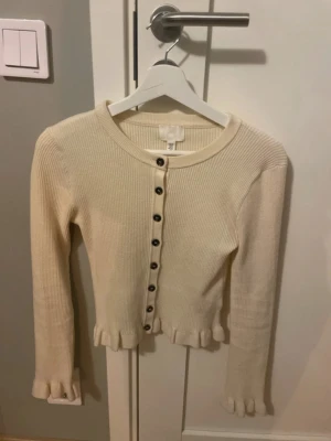 Krämvit ribbad kofta från H&M - Söt krämvit ribbad kofta från H&M med svarta knappar framtill. Koftan har långa ärmar och volangdetaljer vid ärmslut och nederkant. Super populär & söt 🥰