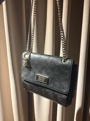 Svart axelväska från Guess - Snygg svart axelväska från Guess med Guess-berlocker.