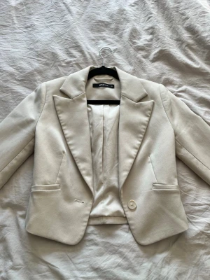 Beige kavaj från Gina Tricot - Stilren beige kavaj från Gina Tricot med klassisk passform, en knapp framtill. Kavajen har två fickor och är helfodrad för en snygg siluett.