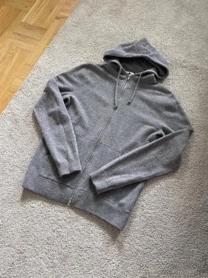 Nn07 Full Zip Hoodie 90% Ull - Säljer en stilren Nn07 No Nationality Ull grå hoodie med dragkedja och huva. Tröjan har ribbade muddar vid ärmslut och nederkant samt fickor framtill. Perfekt för en avslappnad look och enkel att matcha med andra plagg. Materialet känns mjukt och bekvämt.