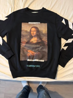 Svart sweatshirt Off-White Mona Lisa - Svart sweatshirt från Off-White med stort Mona Lisa-tryck på framsidan och vita pilar samt texten TEMPERATURE på ryggen. Ärmarna har vita diagonala ränder. Tillverkad i mjuk bomull med rund hals och ribbade muddar. Perfekt för dig som gillar streetwear och konstinspirerad design.