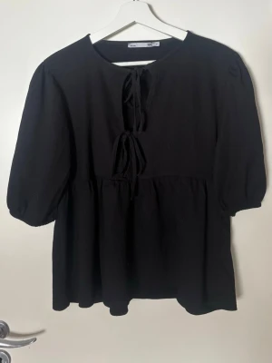 Svart blus med knyt från 157 - Svart blus från 157 med två dekorativa knytband framtill. Blusen har en avslappnad passform, rund halsringning och trekvartsärmar med lätt puff. Materialet är strukturerat och ger en trendig look.