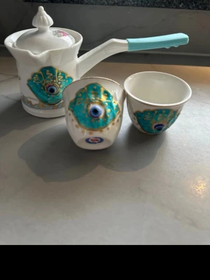 Kaffeset med turkos öga-motiv - Unikt kaffeset i porslin med kanna och två koppar. Setet är vitt med turkosa och guldiga detaljer samt ett dekorativt öga-motiv på både kanna och koppar. Kannan har ett vitt lock och ett handtag med ljusblått grepp. Perfekt för dig som gillar speciella detaljer.