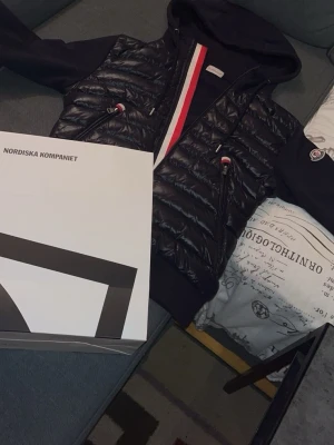 Moncler svart cardiganjacka med huva -  cardigan från Moncler med huva och dragkedja. Jackan har två fickor med dragkedja och detaljer i rött, vitt och blått längs dragkedjan. Priset går att diskutera 