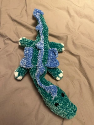 Virkad drakdjur mjukis i blått och grönt - Handgjord mjukis i form av ett drakdjur, virkad i mjuk chenillegarn. Kroppen är grön med blå detaljer längs ryggen och blå vingar. Svansen är lång och slingrig, och tassarna har vita toppar. Svarta ögon i plast ger ett gulligt uttryck. Ca 55 cm lång
