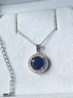 Silverhalsband med blå sten och pärlor i rostfritt stål  - Snyggt halsband i silverfärg med en rund, mörkblå sten i mitten omgiven av små glittrande pärlor. Kedjan är tunn och elegant, och hänget har en klassisk, tidlös design som verkligen sticker ut. Perfekt för dig som gillar stilrena smycken med en touch av färg.