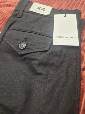 THESE GLORY DAYS T Fairfox Cargo svarta byxor, storlek 44 - Snygga svarta cargobyxor från These Glory Days med raka ben och klassiska cargofickor på sidorna. Byxorna har knappstängning, bälteshällor och är tillverkade i ett slitstarkt bomullstyg. Perfekta för en avslappnad och trendig stil.