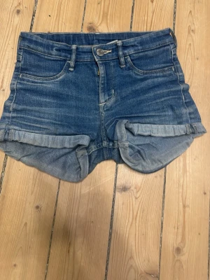 Blå denimshorts med upprullad kant - Snygga blå jeansshorts i klassisk femficksmodell med upprullad kant och knappgylf. Shortsen har en midwaist passform och är tillverkade i bomull för en bekväm känsla. Perfekta för varma sommardagar och enkel att matcha med olika toppar. Storlek 134