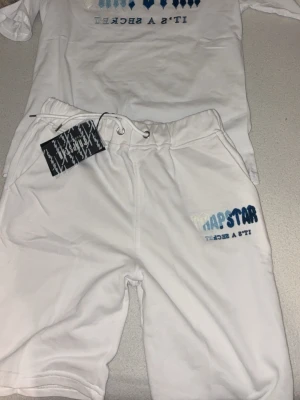 Vit bodysuit-set från Trapstar - Säljer ett vitt bodysuit-set från Trapstar med tryck i blått och ljusblått där det står 'Trapstar' och 'It's a secret'. Setet har en avslappnad passform och är gjort i mjukt bomullsmaterial. Perfekt för dig som gillar streetwear och vill sticka ut.
