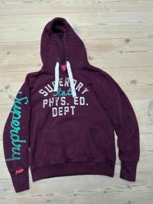 Vinröd hoodie från Superdry - Snygg vinröd hoodie från Superdry med vit och turkos text på bröstet och ärmen. Klassisk känguruficka framtill, vita dragsnören i huvan och ribbade muddar. Perfekt för en avslappnad och sportig stil.