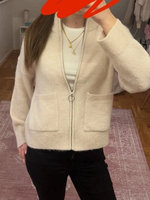 Beige zip-kofta med fickor - Mysig beige kofta med dragkedja och två stora framfickor. Koftan har ribbade muddar och hög krage, perfekt för lager på lager. Materialet ser ut att vara mjukt och fluffigt, troligen ullblandning. Enkel och stilren design som passar till det mesta.