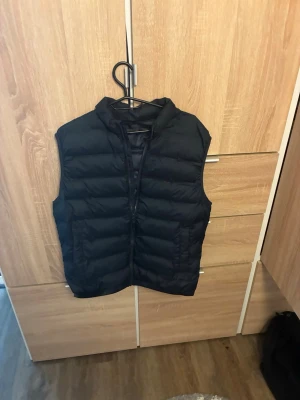 Svart dunväst från ZARA - Snygg svart dunväst från ZARA med quiltad design och hög krage. Västen har dragkedja framtill och två sidofickor. Tillverkad i lätt och glansigt material som ger en modern look. Perfekt att slänga över en hoodie eller tröja för extra värme.
