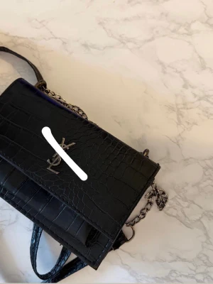 Svart Saint Laurent handväska i croc-mönster - Säljer en svart handväska från Saint Laurent med croc-mönstrad läderlook och silverfärgad kedjerem. Väskan har YSL-logga i metall framtill och en stilren, rektangulär form. Perfekt för dig som gillar lyxiga detaljer och klassisk design.