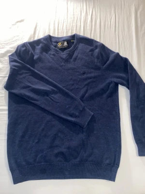 Mörkblå tröja från Lyle & Scott - Snygg mörkblå stickad tröja från Lyle & Scott med rund halsringning och diskret broderad logga på bröstet. Tröjan har ribbade muddar vid ärmslut och nederkant, perfekt för en clean och stilren look.