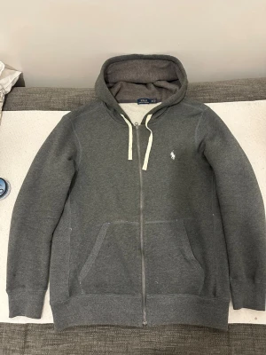 Grå zip hoodie från Polo Ralph Lauren - Mörkgrå hoodie med dragkedja från Polo Ralph Lauren. Klassisk design med vit broderad logga på bröstet, vita snören i huvan och två stora fickor framtill. Tillverkad i mjukt sweatshirtmaterial som är perfekt för en avslappnad stil.