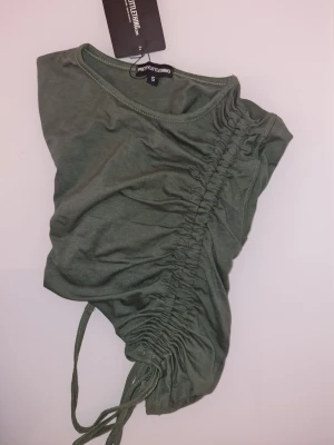 Grön t-shirt med snörning PrettyLittleThing - Grön topp med dragsko längs ena sidan. Dragskon skapar en rynkad effekt och kan justeras för att ändra toppens längd och passform.