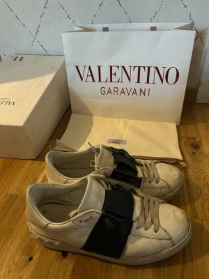 Valentino Garavani vita sneakers - Snygga vita sneakers från Valentino Garavani med bred svart rem över vristen och klassisk låg siluett. Skorna är tillverkade i läder och har diskreta detaljer samt logga på plösen. Perfekta för dig som gillar exklusiv streetstyle.