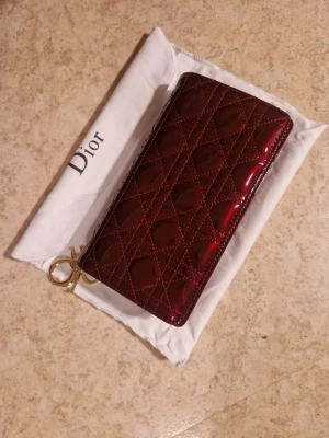 Röd lackad plånbok från Dior - Snygg röd plånbok från Dior i glansigt lackat material med quiltat mönster och guldfärgad dragkedja. Kommer med original dustbag. Perfekt accessoar för dig som gillar lyxiga detaljer och klassisk design.