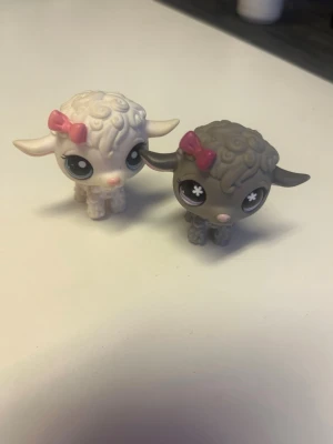 Littlest Pet Shop lammfigurer - Två söta samlarfigurer från Littlest Pet Shop föreställande lamm. Ena lammet är vitt med rosa rosett och stora ögon, det andra är grått med rosa rosett och vita detaljer i ögonen. Båda är tillverkade i plast och har gulliga lockiga detaljer på huvudet.