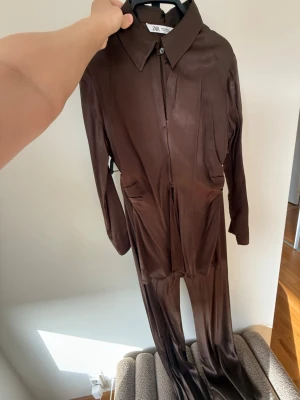 Brun över och underdel från Zara med krage - Säljer en brun långärmad överdel från Zara med klassisk krage och knappar framtill. Byxorna till har vida ben,Materialet är glansigt och ger en cool look (viskos). Perfekt för dig som vill sticka ut med en trendig och bekväm outfit.