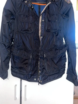 Parajumper windbreaker  - Snygg mörkblå vindjacka från Parajumpers med flera fickor och parajumper märke. Jackan har dragkedja, knappar och justerbar midja. Materialet är glansigt och lätt, perfekt för att skydda mot vind. Det finns en inbyggd luva du kan ta ut om du vill. 