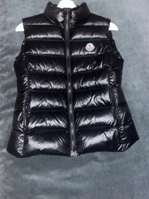 Svart dunväst från Moncler - Snygg svart dunväst från Moncler med glansig finish och hög krage. Västen har en klassisk quiltad design, dragkedja framtill och Moncler-logga på bröstet. Perfekt för lager-på-lager och ger en trendig look till din outfit.