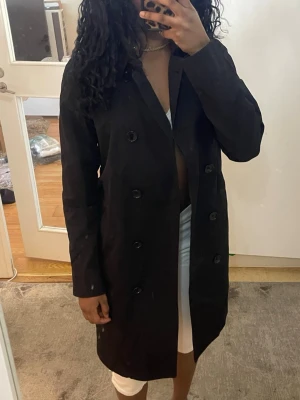 Svart klassisk trenchcoat - Snygg svart trenchcoat med dubbelknäppning och raka ärmar. Jackan har en tidlös design med knappar framtill och en rak passform som ger en clean look. Perfekt för dig som vill ha en stilren och enkel trenchcoat till garderoben.