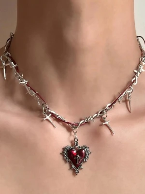 Halsband med rött hjärthänge och taggar - Unikt halsband med silverfärgade taggar och ett stort rött hjärthänge i metall. Kedjan har detaljer som påminner om taggtråd och små korshängen. Perfekt för dig som gillar alternativ stil och vill sticka ut med accessoarer i metall och röd emalj.