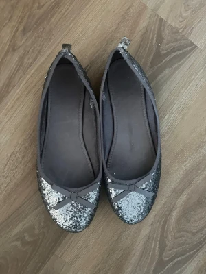 Glittriga grå ballerinaskor med rosett - Säljer ett par glittriga grå ballerinaskor från H&M i storlek 38. Tyvärr har de gått sönder lite i hälen (se bild 3) men de funkar bra ändå!