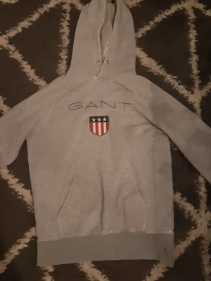 Grå hoodie från GANT med logga - Snygg grå hoodie från GANT med stor broderad logga och sköld på bröstet. Klassisk känguruficka framtill och justerbar huva med metallöljetter. Tillverkad i mjuk bomullsblandning för extra komfort. Perfekt för en avslappnad och sportig stil.