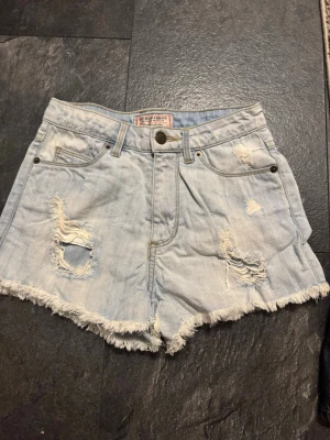 Ljusblå denimshorts från Guess - Kollar intresset! Säljs vid bra pris!  ljusblå denimshorts från Guess med slitna detaljer och fransig kant. Shortsen har klassisk femficksdesign, hög midja och ett broderat Guess-märke på bakfickan. Perfekta för sommaren och riktigt snygga till sneakers eller sandaler.