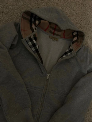 Grå hoodie från Burberry - Snygg grå hoodie från Burberry med dragkedja och klassiskt rutigt foder i huvan. Hoodien har ribbade muddar, fickor framtill och är tillverkad i mjukt bomullsmaterial. Perfekt för en avslappnad men stilren look.