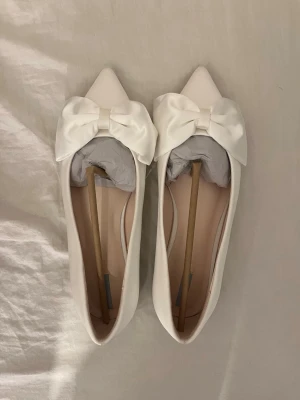 Vita ballerinaskor med rosett - Stilrena vita ballerinaskor från Next med spetsig tå och stor, elegant rosett i satin framtill. Skorna har en slät ovandel och låg klack, vilket ger en klassisk och chic look. Perfekta för dig som gillar en feminin och trendig stil.