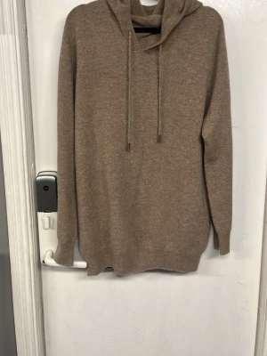Beige hoodie i mjukt material - Säljer en stilren beige hoodie med huva och dragsnören. Hoodien har långa ärmar och ribbade muddar vid ärmslut och nederkant. Materialet känns mjukt och skönt, perfekt för en avslappnad look. Märke saknas på plagget.