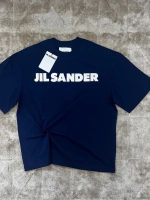 Marinblå Jil Sander t-shirt - Jil Sander t-shirt. Helt ny med tags kvar. Tillverkad i mjuk bomull med en premium känsla och ren finish. Klassisk passform som sitter snyggt utan att vara för tight. Enkel design med tydlig logga som ger en stilren look. Väldigt lätt att matcha och funkar i princip till allt. Skick 10/10. Vid fler frågor gärna kontakta mig privat. Priset är förhandlingsbart!