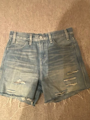 Blå jeansshorts med slitningar - Säljer ett par ljusblå jeansshorts med slitna detaljer och fransig kant. Shortsen har klassisk femficksdesign, bälteshällor och knappgylf. Perfekta för sommaren och ger en avslappnad streetstil.