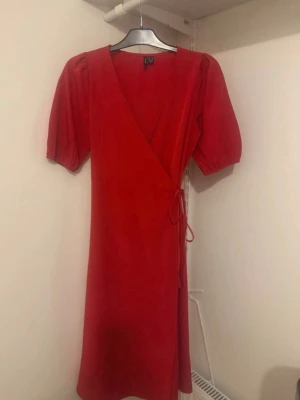 Röd omlottklänning med puffärm - Snygg röd omlottklänning med puffärmar och knyt i midjan. Klänningen har v-ringning och är tillverkad i ett mjukt tyg som ger en skön känsla. Perfekt för dig som vill ha en enkel men trendig look.