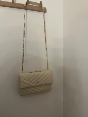 Beige quiltad axelväska med kedja - Snygg beige axelväska i quiltat mönster med guldfärgad kedjerem och liten gulddetalj framtill. Väskan har en klassisk rektangulär form och stängs med lock. Perfekt för dig som gillar stilrena accessoarer. Nypris 400kr. Pris kan diskuteras!🤍 