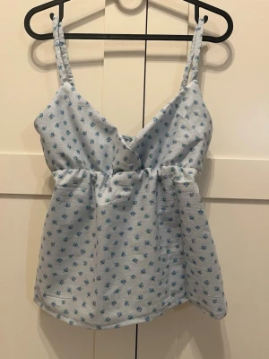 Ljusblå blommig linnetopp - Supersöt ljusblå linnetopp med små blå blommor och smala axelband. Toppen har en omlott-v-ringning och resår i midjan som ger en fin passform. Perfekt för varma dagar och enkel att matcha med jeans eller shorts.