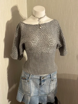 Grå stickad topp med spetsmönster - Snygg grå stickad topp med bred ribbad midja och korta, vida ärmar. Överdelen har ett detaljerat spetsmönster i geometriska former och båtringad hals. Perfekt för dig som gillar en unik och trendig look. Materialet känns mjukt och stretchigt.