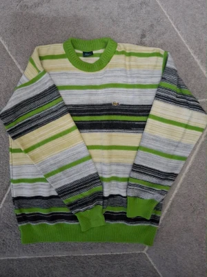 Randig stickad tröja från Lacoste - Snygg stickad tröja från Lacoste med breda ränder i grönt, grått, svart och beige. Tröjan har rund halsringning och ribbade muddar i grönt. Klassisk Lacoste-logga med krokodil på bröstet. Perfekt för dig som gillar retrovibbar och färg.