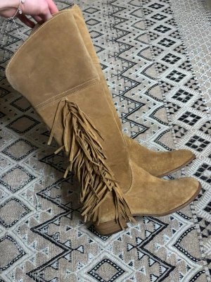 Beige mocka cowboy boots med fransar - Snygga beige cowboy boots i mocka med lågt skaft och coola fransdetaljer längs sidan. Bootsens spetsiga tå och låga klack ger en klassisk westernkänsla. Dragkedja på insidan för enkel påtagning. Perfekta för dig som vill sticka ut. Eller piffa upp en outfit.