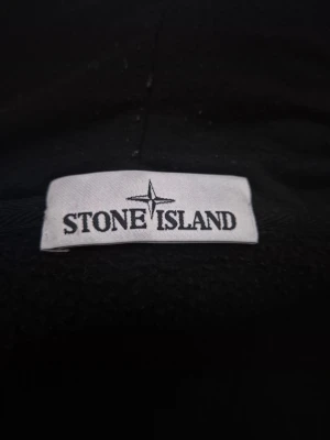 Svart hoodie från Stone Island - Snygg mörkgrön hoodie från Stone Island med dragkedja framtill, justerbar huva med snören och klassisk känguruficka. Märkeslapp på insidan och detalj på ärmen. Perfekt för dig som gillar streetwear och vill ha något stilrent och bekvämt.