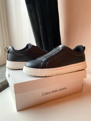 Svarta Calvin Klein Jeans sneakers - Stilrena svarta sneakers från Calvin Klein Jeans med präglad CK-logga på sidan och vit tjock sula. Skorna är låga och har en clean design i slätt lädermaterial. Perfekta för dig som gillar minimalistisk och modern stil. Ny pris 1000kr dem är helt nya bara använda en gång storlek 39 men kommer stor i storlek så passar även 40 dem är unisex alltså killar och tjejer kan ha på sig dem     Pris kan diskutera 