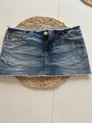 Blå jeanskjol med rå kant 36 midjemått  - Snygg blå jeanskjol i kort modell med rå, fransig kant nertill. Kjolen har klassiska fem fickor, knapp- och dragkedjestängning samt slitna detaljer för en trendig look. Perfekt att matcha med en enkel t-shirt eller hoodie.