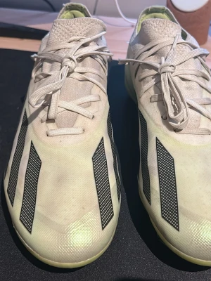 Adidas vita fotbollsskor med svarta ränder - Säljer ett par snygga vita Adidas fotbollsskor med klassiska svarta ränder och gulaktig sula. Skorna har snörning och är tillverkade i syntetmaterial, perfekta för dig som vill ha stilrena och lätta skor på planen.