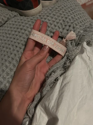 Beige broderat Dior armband - Säljer ett beige tygarmband från Christian Dior med texten 'Christian Dior J'Adior' broderat i vitt. Armbandet har dekorativa tofsar i ändarna och knyts runt handleden. Perfekt accessoar för dig som gillar exklusiva detaljer och trendig design.